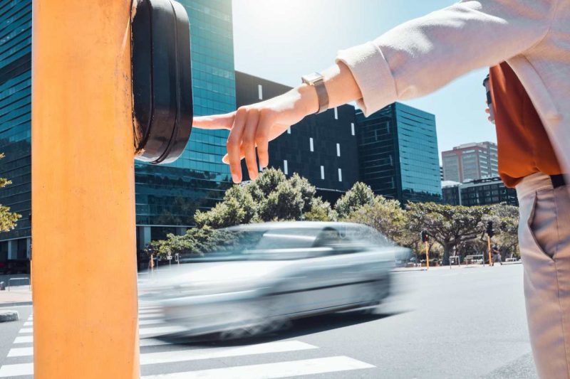 A pedestrian hits a crosswalk signal while a car drives past on a city road.