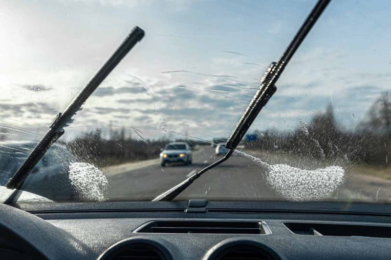 A car's windshield wipers clear debris off a windshield on a sunny day.