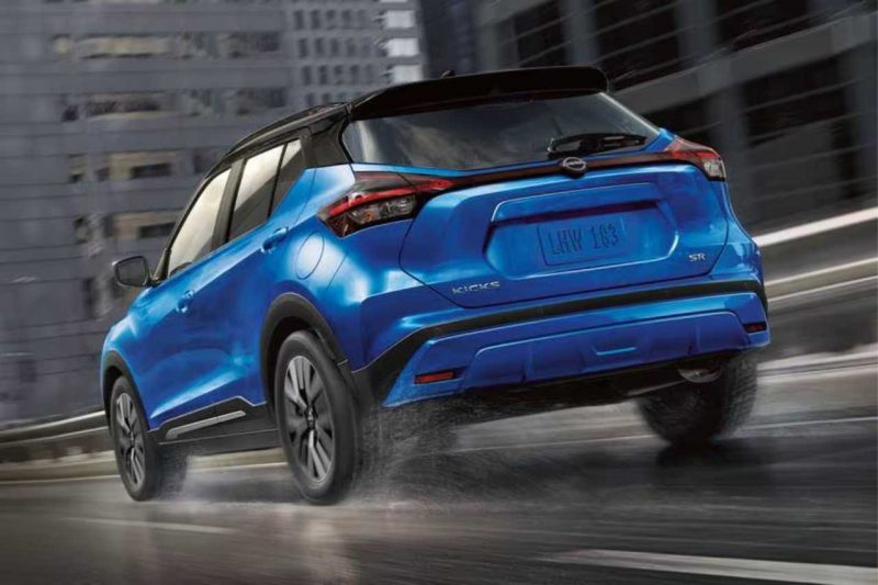 The rear exterior of a blue 2024 Nissan Kicks SUV driving on a wet, rainy city road.
