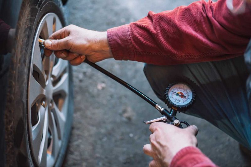 A man in a red shirt performing car maintenance and checking his tire pressure with a gauge.