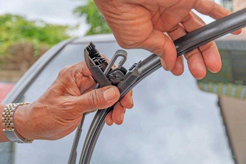 A man replacing his windshield wiper with a hook connector, performing car maintenance on a summer day.