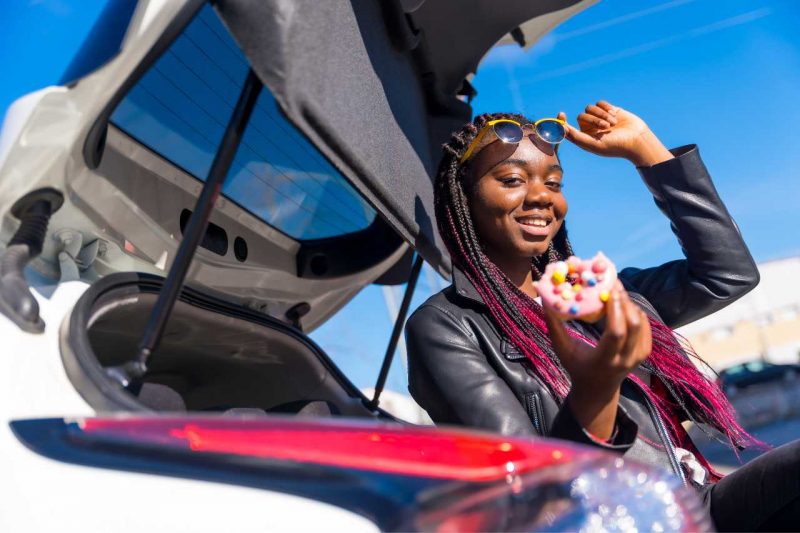 An African American woman poses in front of a car with a pink donut.