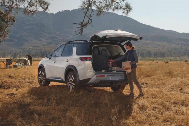 A Caucasian woman uses the motion-activated liftgate feature to load items into the back of her Nissan Pathfinder. The vehicle is parked in a field on a camping trip.