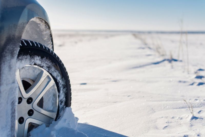 A vehicle wearing winter tires drives through the snow on a sunny day.