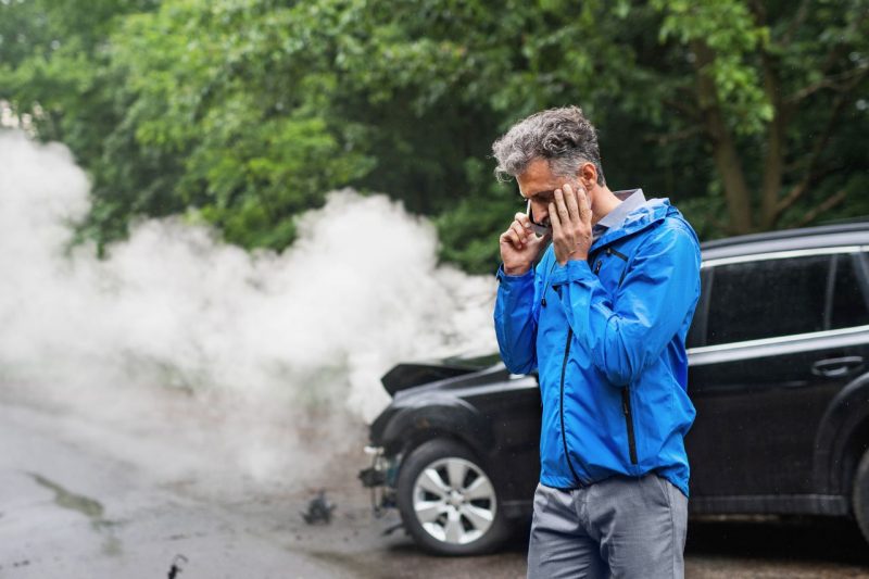A middled aged Caucasian man in a blue jacket stands in front of his broken down car, which has smoke coming from the hood.