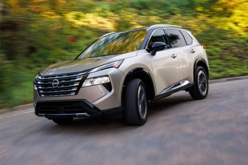 A champagne coloured 2025 Nissan Rogue SUV is driving down a sunny road with greenery in the background.
