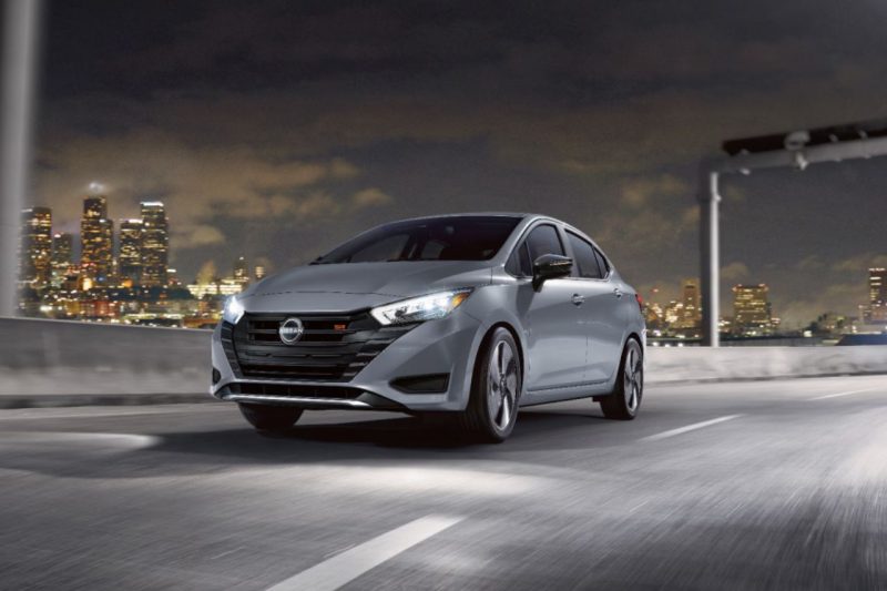 A grey Nissan sedan drives down a lit-up highway at night with city lights in the background.