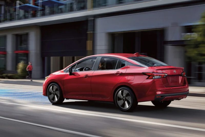 A red 2025 Nissan Versa sedan drives down city roads, utilizing safety and driver technologies.
