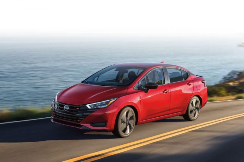 The 2025 Nissan Versa in a red colour drives down a sunny highway next to a body of water.