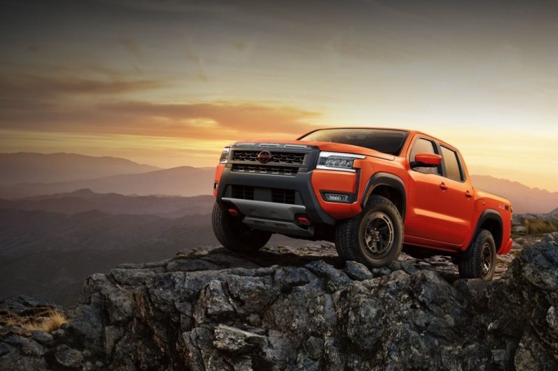 An orange Nissan Frontier truck stands atop a mountain in front of a sunset.