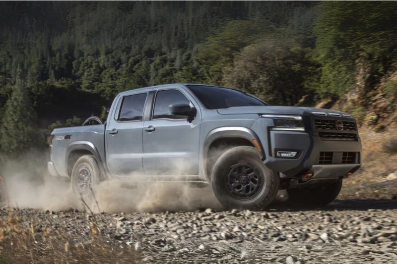 A dusty blue-grey Nissan Frontier truck spins its wheels in a dusty river bed.
