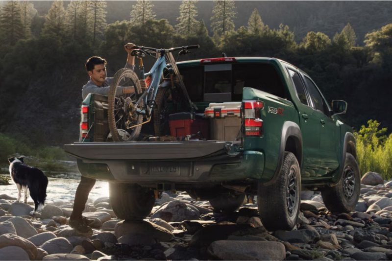 A man loads bikes and gear into the truck bed of his 2025 Nissan Frontier, with a dog and lake in the background.